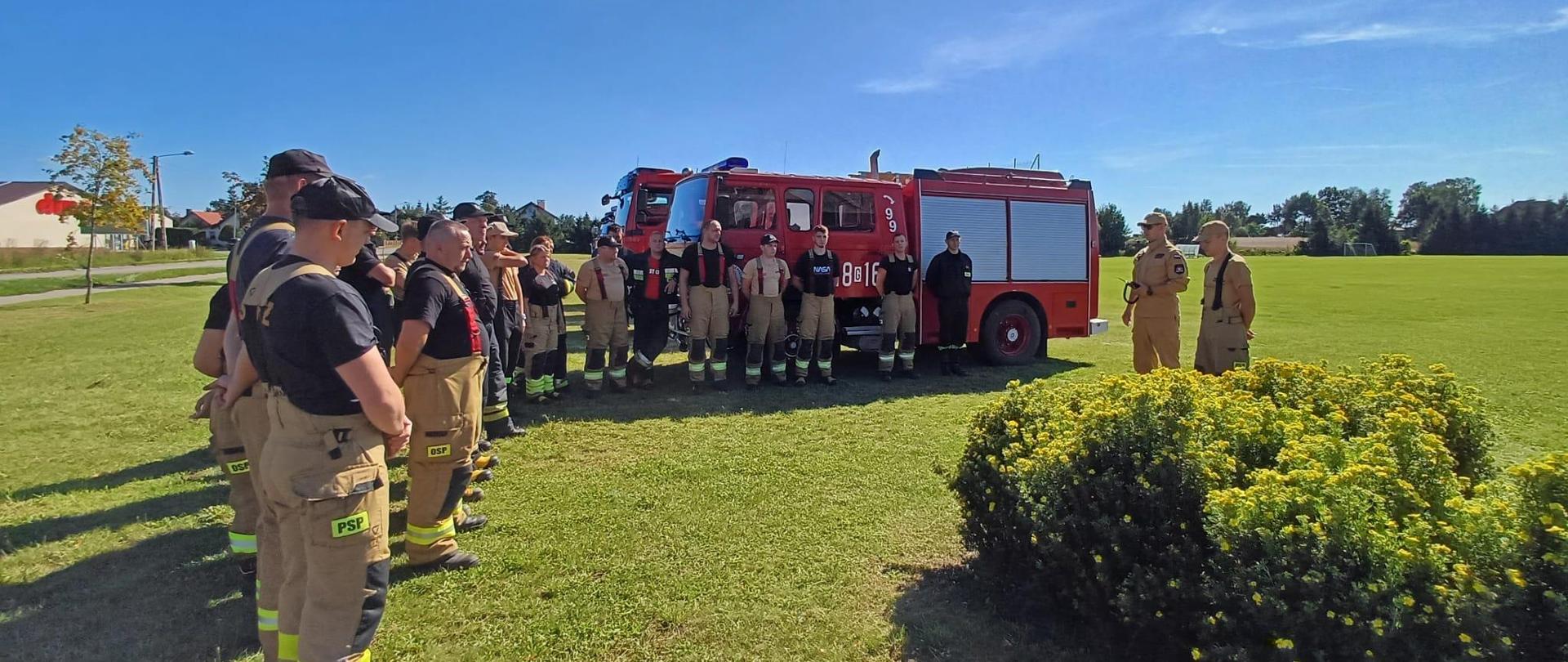 Słoneczny dzień, odprawa przed ćwczeniami. Dowódcy ćwiczeń stoją przed uczestnikami ćwiczeń na polanie. W tle wozy strażackie.