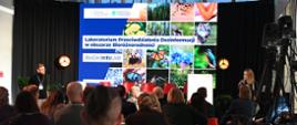 Na zdjęciu widać publiczność siedzącą na krzesłach tyłem. Publiczność patrzy na ekran przed sobą, na którym widnieje napis "Laboratorium przeciwdziałania dezinformacji w obszarze bioróżnorodności". Na scenie przy ekranie stoi również kobieta z mikrofonem oraz mężczyzna.