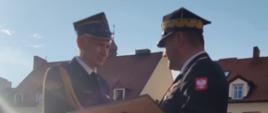 Komendant Wojewódzki wręcza Komendantowi Miejskiemu Dyplom Uznania. W tle poczty sztandarowe oraz kamienice. 