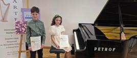 Zdjęcie przedstawia dwóch uczniów po lewej stronie stoi chłopiec trzymajacy dyplom obok na stołku siedzi dziewczynka w sukience również trzyma dyplom. Po prawej stronie stoi czarny fortepian.