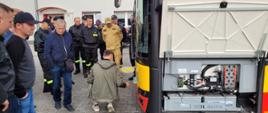 Przód autobusu stojący na utwardzonym placu z otwartą klapą serwisową, który oglądają zgromadzeni strażacy i osoby cywilne 