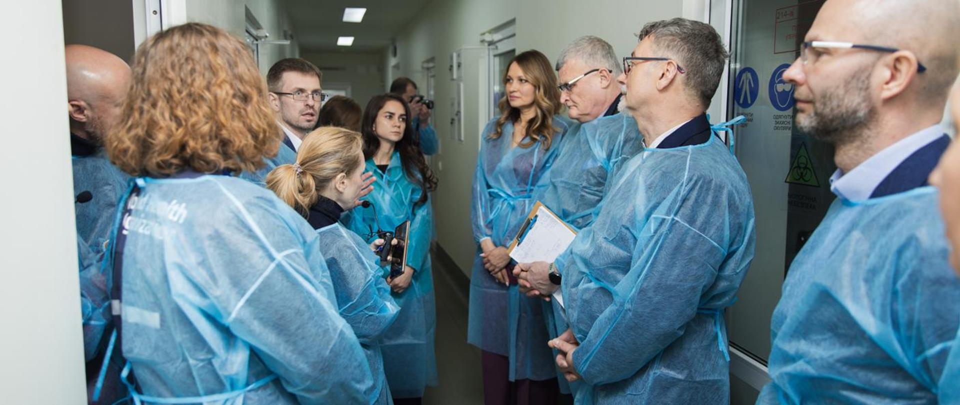 Przedstawiciele Głównego Inspektoratu Sanitarnego wraz z Głównym Inspektorem Sanitarnym Pawłem Grzesiowskim stojący na korytarzu w budynku w niebieskich fartuchach