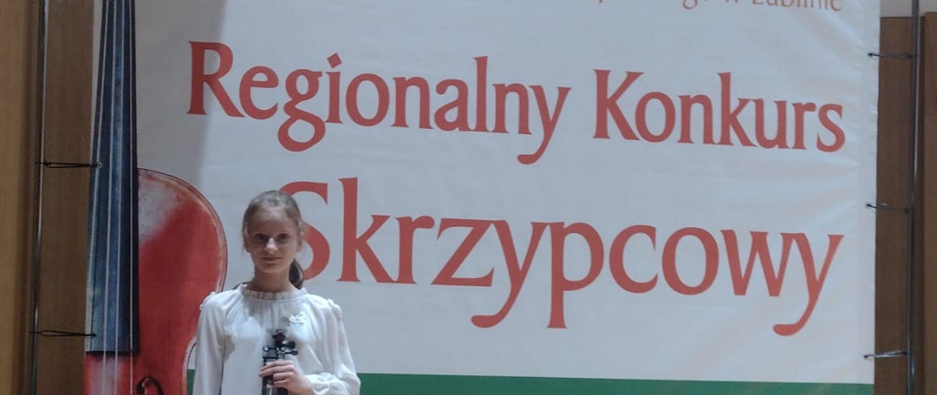 Na zdjęciu znajduje sie dziewczynka ze skrzypcami która stoi na scenie. w tle jest duzy plakat z napisem XVIII Regionalny Konkurs Skrzypcowy „Skrzypcowe spotkania”