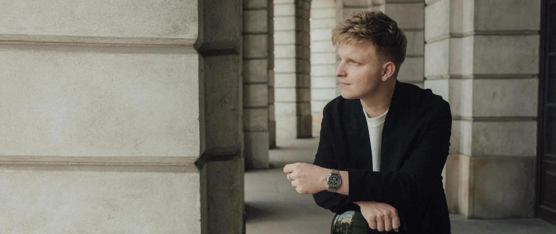 Zdjęcie gitarzysty z czarnym futerałem od instrumentu, ubranym w białą koszulkę i czarną marynarkę na tle białych, kamiennych wnętrz