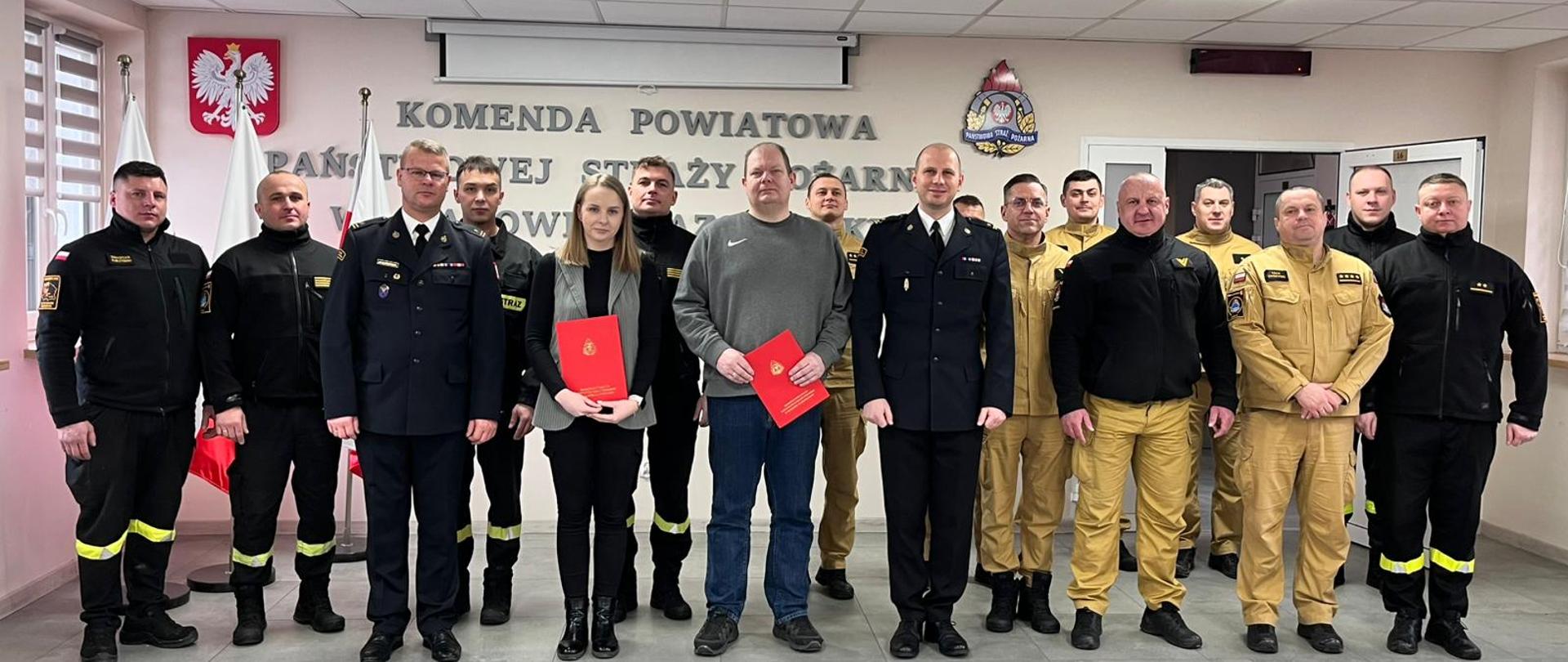 Wspólne zdjęcie pracowników cywilnych z funkcjonariuszami Komendy Powiatowej PSP. Komendant wraz z Zastępcą ubrani są w mundury wyjściowe, funkcjonariusze zmianowi w czarne ubrania a funkcjonariusze systemu codziennego w stroje w kolorze piaskowym