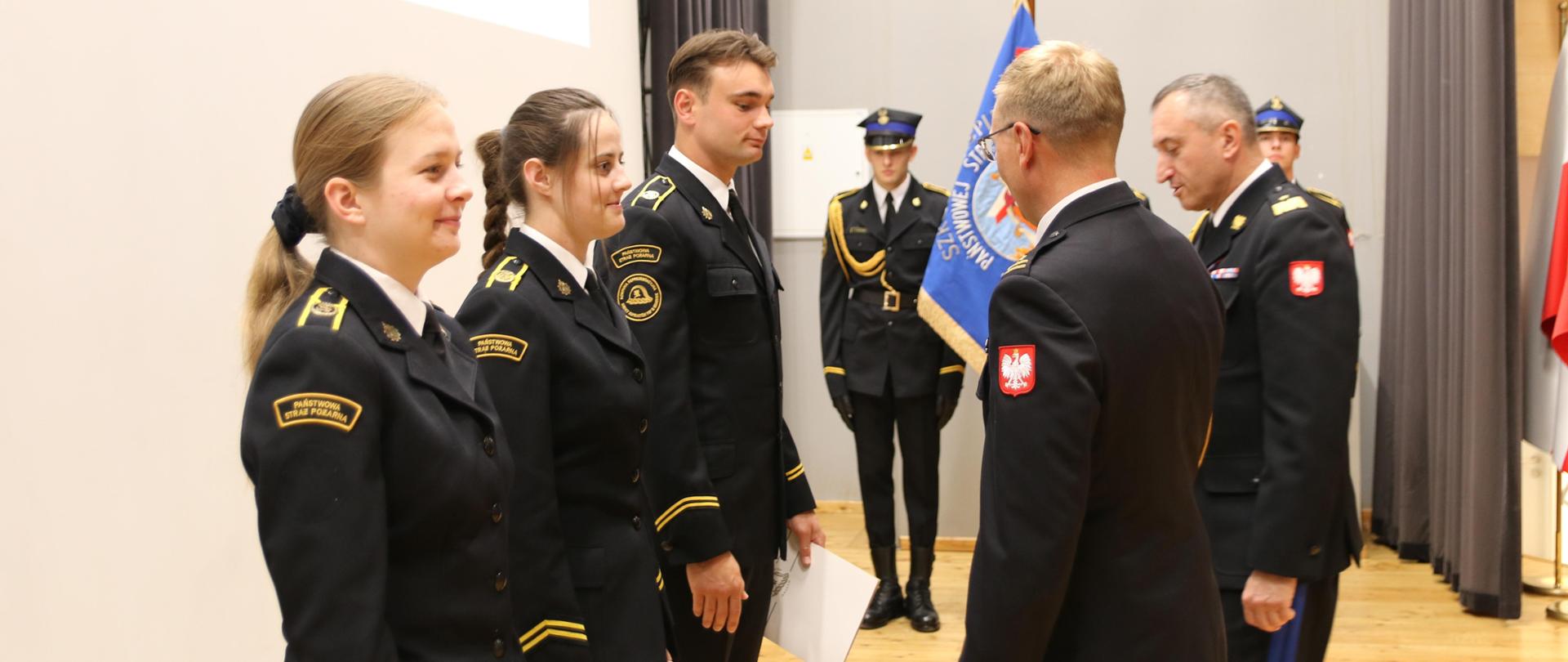 Scena auli SA PSP w Krakowie. Wręczanie teczek z dokumentami w trakcie uroczystego zakończania roku szkolnego. Strażacy w wyjściowych mundurach. 