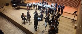 Grająca orkiestra smyczkowa, Pani Dyrygent, za nimi śpiewający Chór PSM na scenie sali koncertowej PSM