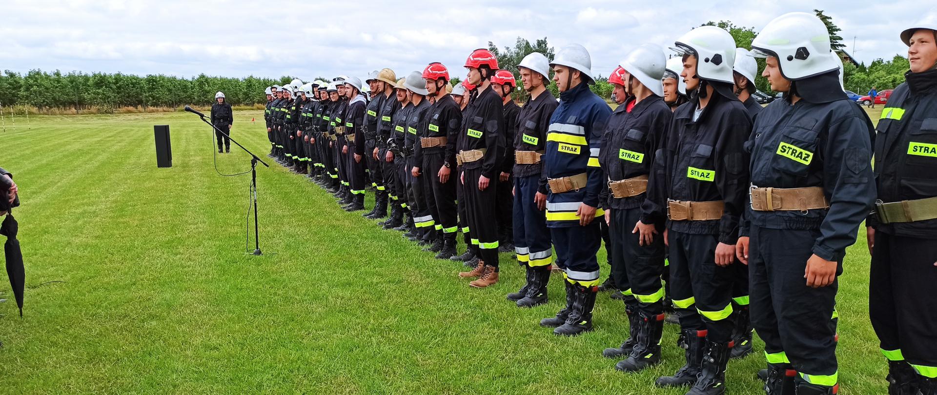 Zdjęcie przedstawia strażaków OSP ustawionych po prawej stronie w dwuszeregu. Ubrani są w ubranie koszarowe, pasy strażackie oraz hełmy. Strażacy stoją na boisku z trawą.