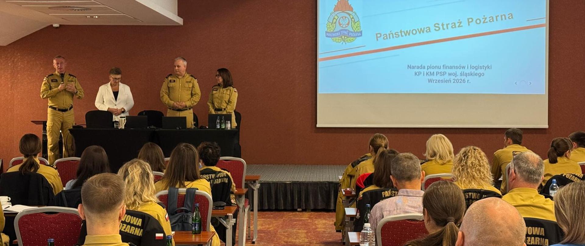 Zdjęcie przedstawia naradę służbową odbywającą się w sali konferencyjnej. Z przodu, przed stołem prezydialnym na niewielkim podwyższeniu, stoi czworo prelegentów: trzech funkcjonariuszy Państwowej Straży Pożarnej w piaskowych mundurach służbowych oraz kobieta w białej żakiecie. Po prawej stronie wisi duży ekran projekcyjny z prezentacją opatrzoną logo PSP oraz napisem informującym o naradzie pionu finansów i logistyki (wrzesień 2026 r.). Na pierwszym planie widać widownię siedzącą przy rzędach stołów. Większość uczestników to strażacy w piaskowych mundurach służbowych.