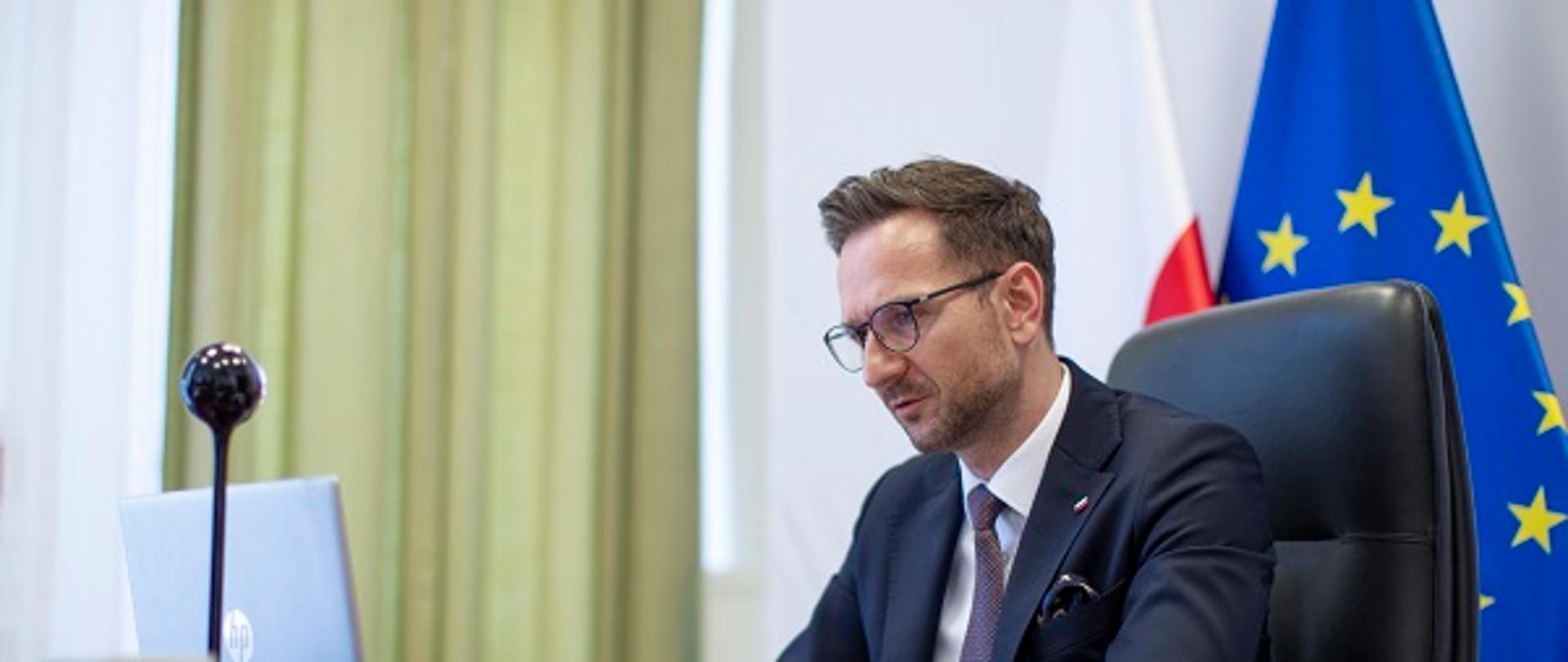 Minister Waldemar Buda siedzi przy biurku przed laptopem. Z tyłu flagi Polski i UE.
