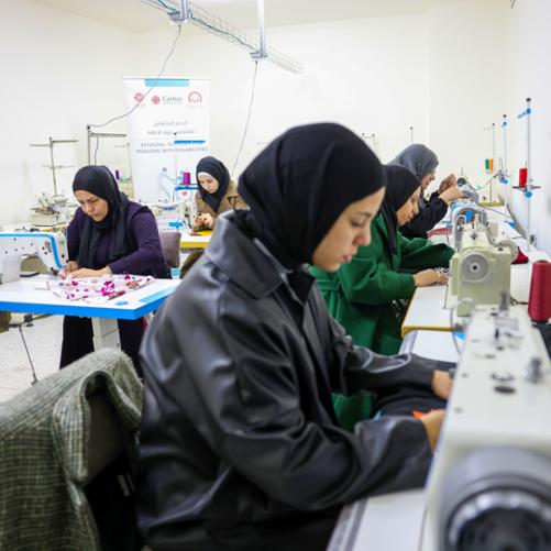 5 kobiet szyje na maszynach, z tyłu widać banner z logo Polska pomoc i Caritas Jerusalem