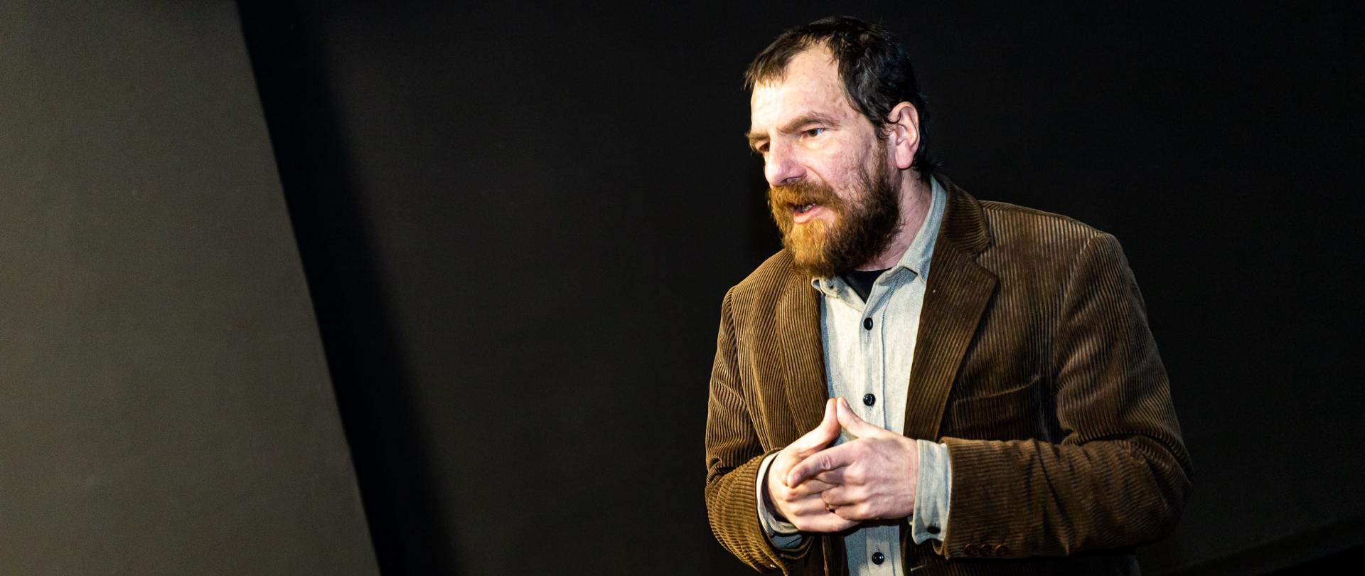 Zdjęcie przedstawia postać profesora habilitowanego Krzysztofa Moraczewskiego, dorosłego mężczyzny w marynarce, z brodą, który podczas wykładu stoi w ciemnej sali teatralnej z rękoma złożonymi w jednym kierunku. 