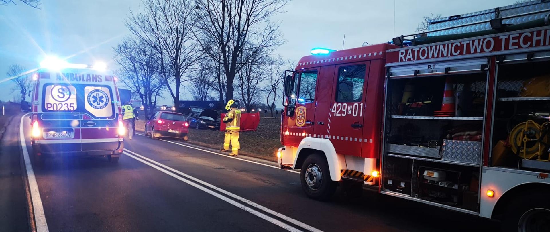 Po lewej stronie zdjęcia widać karetkę pogotowia. Jest godzina wieczorna lekki półmrok. Po prawej stronie widać samochód strażacki przed samochodem stoi strażak w mundurze koloru piaskowego. W tle po prawej stronie widać drzewa a za nimi na polu stoi samochód. Przed samochodem rozwinięty jest czerwony strażacki parawan