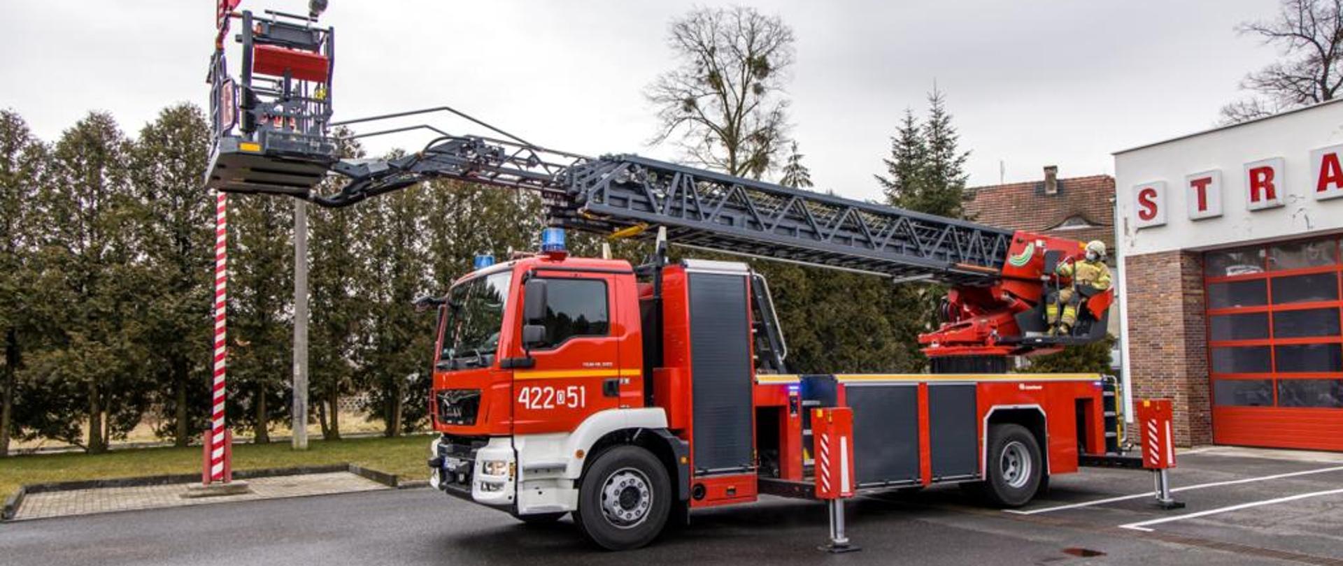 Samochód pożarniczy drabina a w tle drzewa i budynek strażnicy