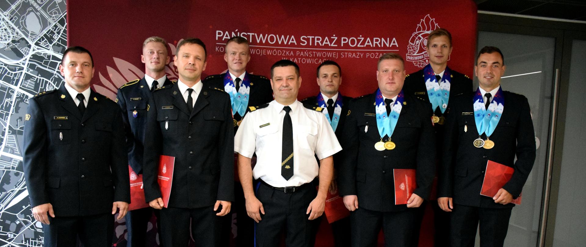 Na zdjęciu ścianka w kolorze czerwonym z godłem Polski, po lewej stronie, u góry napis Państwowa Straż Pożarna Komenda Wojewódzka Państwowej Straży Pożarnej w Katowicach. W prawym górnym rogu logo Państwowej Straży Pożarnej. Na tle ścianki stoją: po środku Śląski Komendant Wojewódzki PSP nadbryg. Jacek Kleszczewski w białej koszuli i czarnych spodniach z lampasami. Obok niego siedmiu wyróżnionych strażaków - sportowców w czarnych mundurach wyjściowych. Pięciu z nich ma na szyjach medale, które zdobyli na Światowych Igrzyskach Służb Mundurowych. Po lewej stronie stoi w mundurze wyjściowym naczelnik Ośrodka Szkolenia Komendy Wojewódzkiej PSP w Katowicach st. kpt. Adam Gawron.