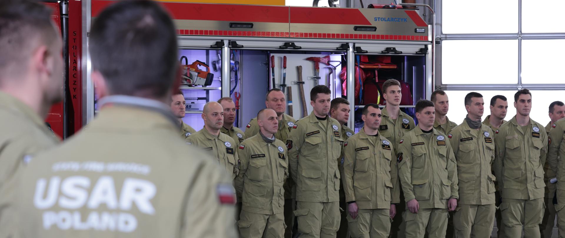 strażacy stoją w szeregu , jeden z nich tyłem z napisem na mundurze USAR POLAND