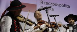 Konferencja Fundusze Europejskie dla kultury, gra kapela góralska Beskid, w środku siedzi kobieta ze skrzypcami ubrana w góralski strój ludowy, po prawej akordeonista, a po lewej skrzypek także ubrani w góralskie stroje ludowe