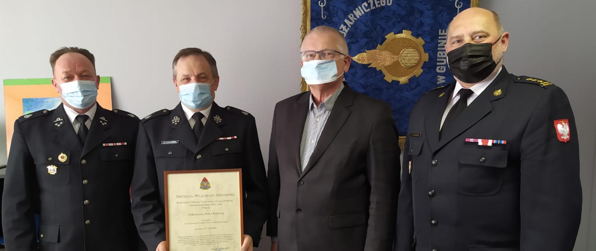 Zdjęcie przedstawia stojących w szeregu od lewej dh Jarosława Benc, dh Ryszarda Kasowskiego, Jana Wiśniewskiego oraz st. bryg. Andrzeja Kaźmieraka. Dh Ryszard Kasowski trzyma w dłoniach Decyzje Komendanta Głównego PSP w sprawie włączenia jednostki Ochotniczej Straży Pożarnej w Połęcku do Krajowego Systemu Ratowniczo-Gaśniczego.