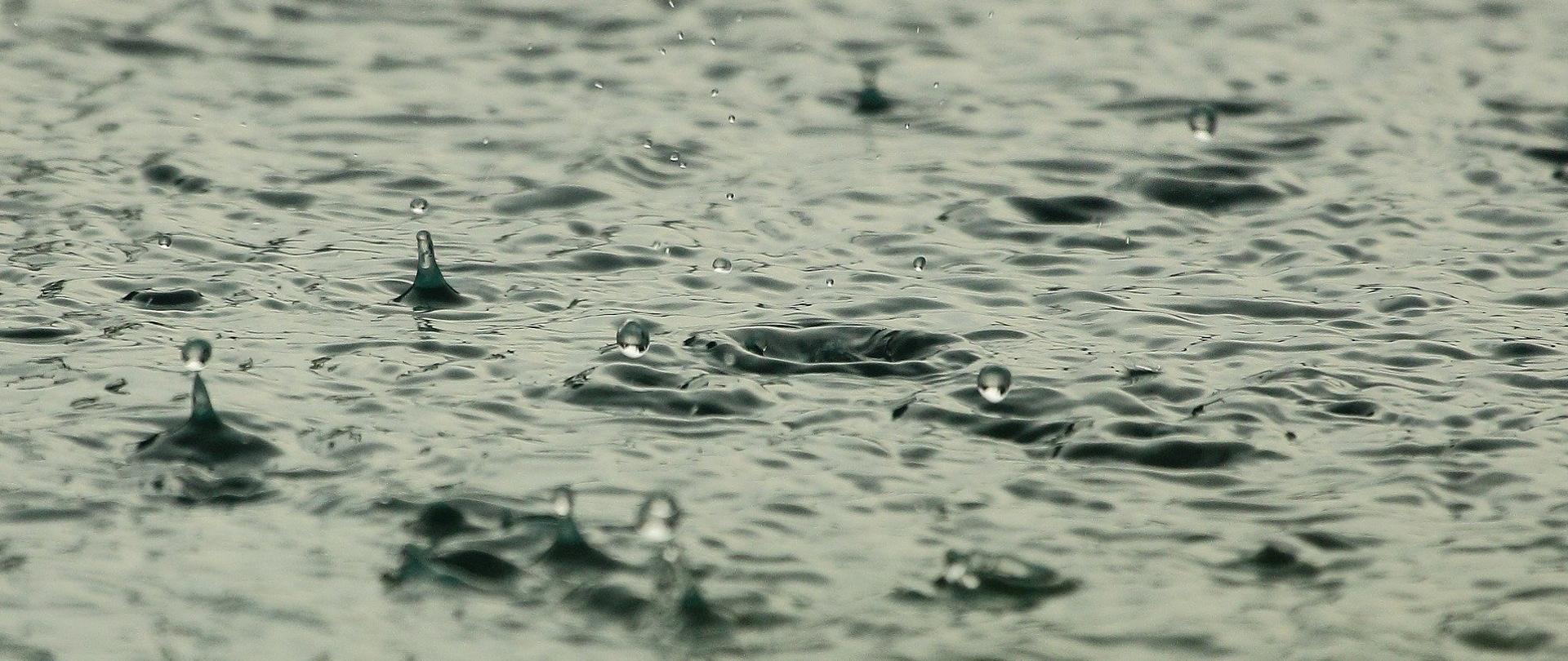 Zdjęcie przedstawia taflę wody i uderzające o nią duże krople deszczu. Całość utrzymana w kolorystyce sepii. 