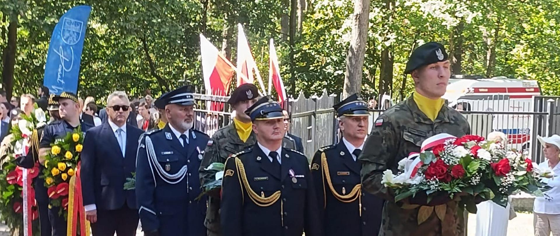 Przedstawiciele służb mundurowych oraz delegacje z wieńcami podczas uroczystości rocznicowych na terenie leśnym.