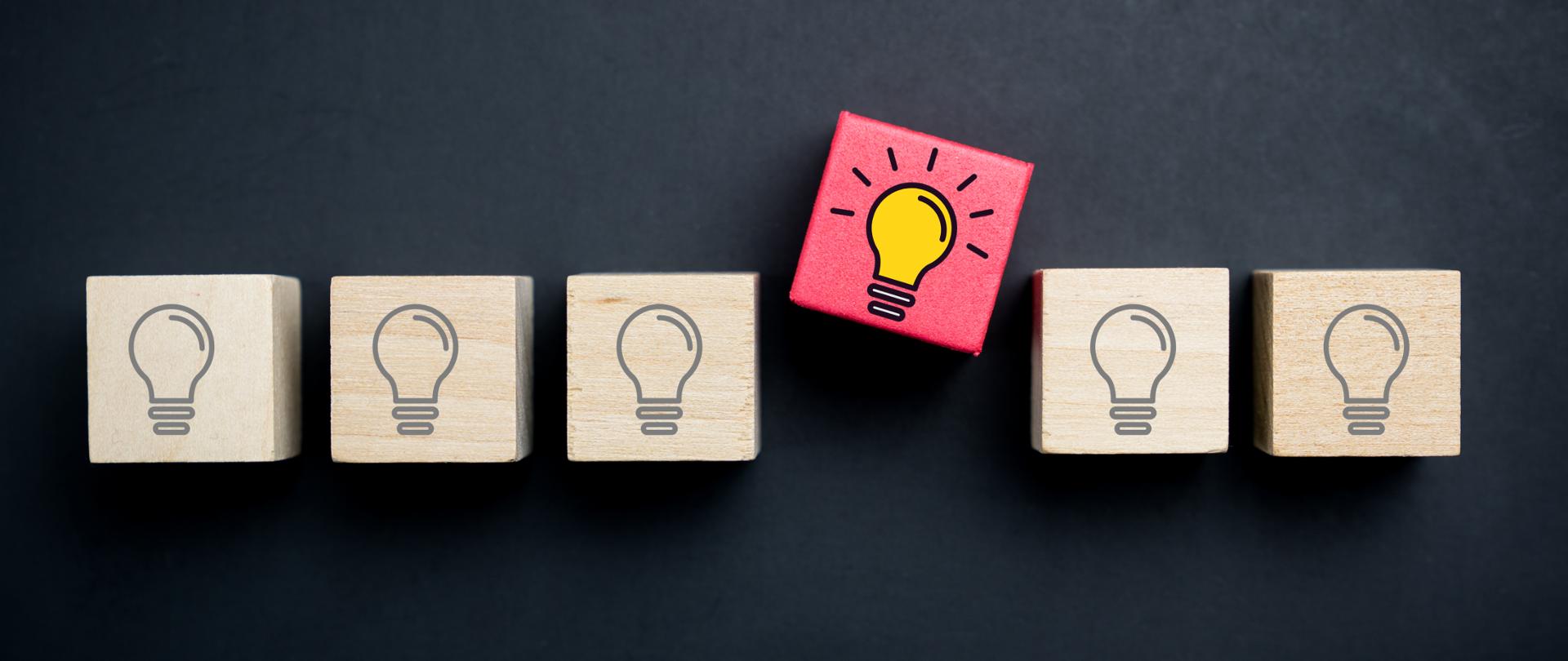 Grafika z czarnym tłem. Obrazek przedstawia drewniane klocki, na których narysowane są żarówki. Jeden klocek (wysunięty do góry) jest kolorowy, a żarówka zapalona.