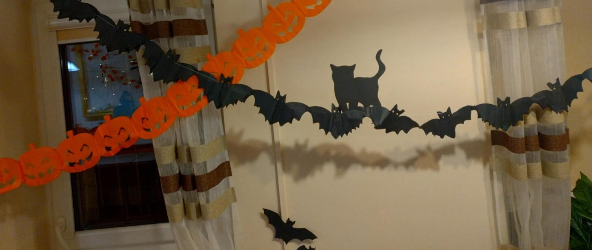 Ręczne ozdoby jedne z nich to czarny papierowy łańcuch wycięty w formie nietoperzy, po środku z czarnym kotem, druga ozdoba to papierowy łańcuch w kształcie pomarańczowych dyń.