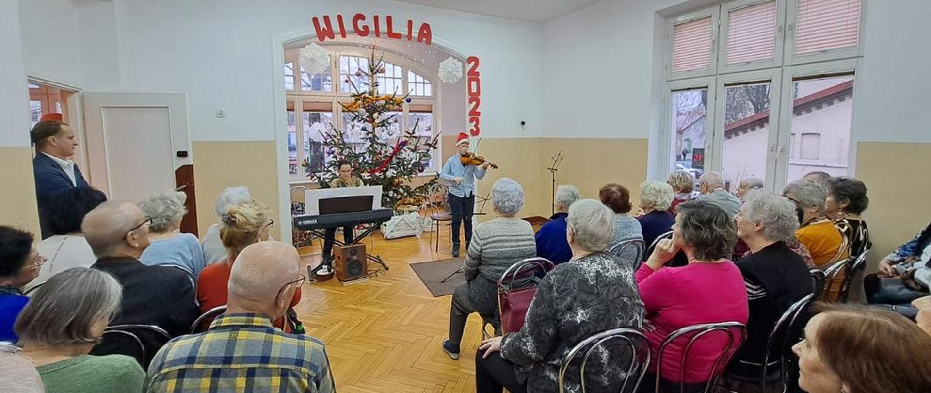 Zdjęcie kolorowe. Seniorzy słuchają koncertu na skrzypce i fortepian. W tle młody skrzypek i pianistka przy swoich instrumentach. W oddali choinka.