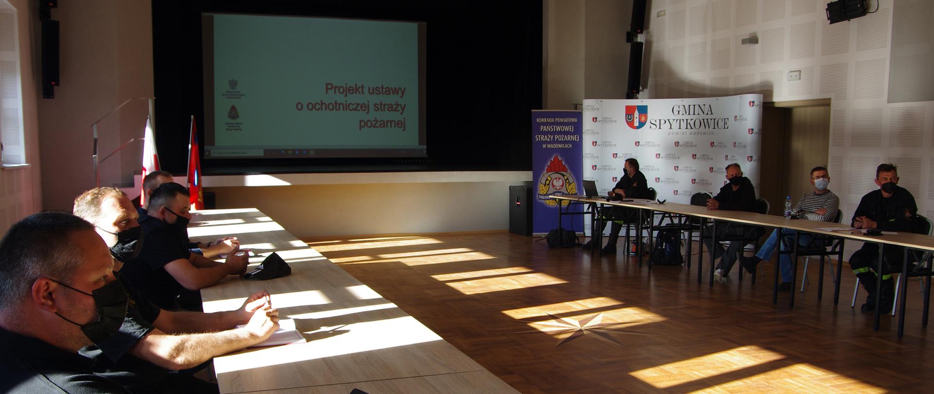Widok ogólny sali sesyjnej. Na pierwszym planie członkowie OSP w maseczkach ochronnych siedzą przy stołach. Na drugim planie ekran z wyświetlonym projektem ustawy o OSP. W tle po prawej stronie widoczny baner z logo KP PSP Wadowice oraz tzw. ścianka z logo Gminy Spytkowice, po lewej stronie stojak z flagą Polski i flagą Związku OSP RP. 