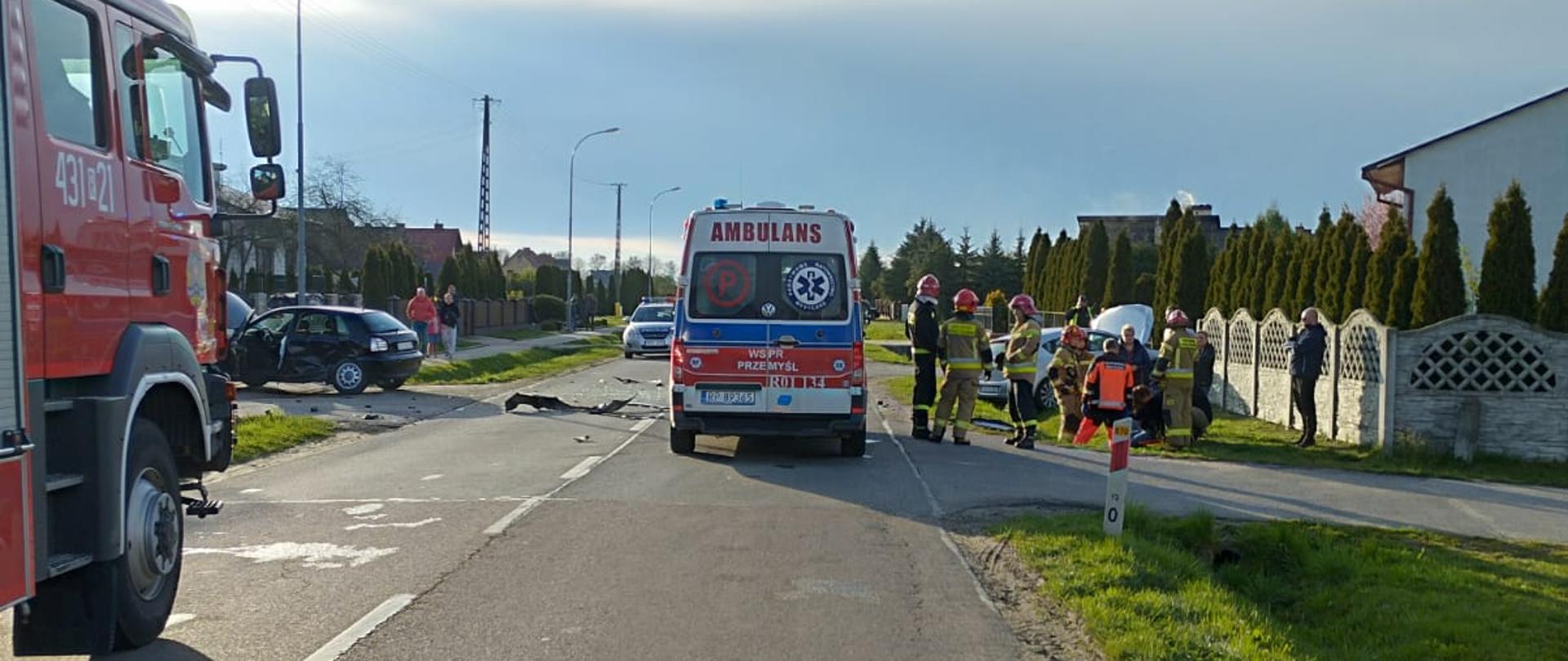 Na zdjęciu widzimy scenę z wypadku samochodowego na drodze wojewódzkiej. Po lewej stronie widać częściowo wóz strażacki, na środku ambulans, a z prawej strony siedzącą na rowie osobie poszkodowaną, której pomocy udzielają strażacy i ratownicy medyczni.