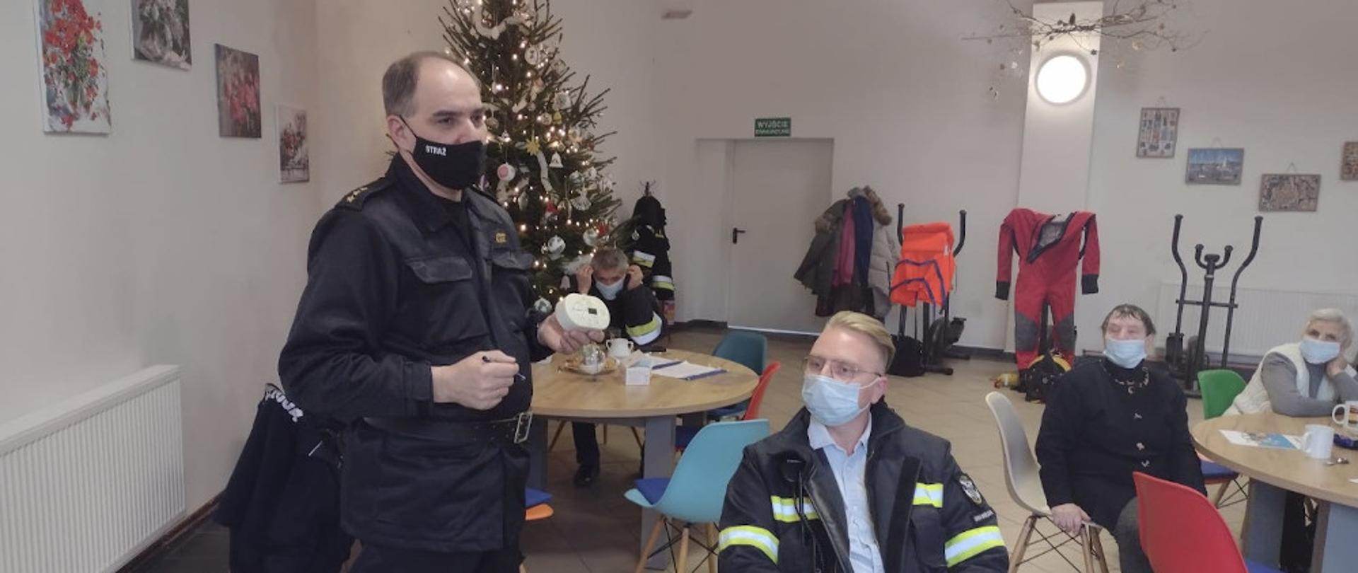 Przedstawiciel Komendy Powiatowej PSP w Szczecinku kapitan Mirosław Śledź, ubrany w ubranie dowódczo sztabowe, koloru czarnego trzymając w lewym ręku czujkę tlenku węgla prezentuje urządzenie i omawia jak powstaje tlenek węgla i jak zapobiegać jego powstawaniu. 