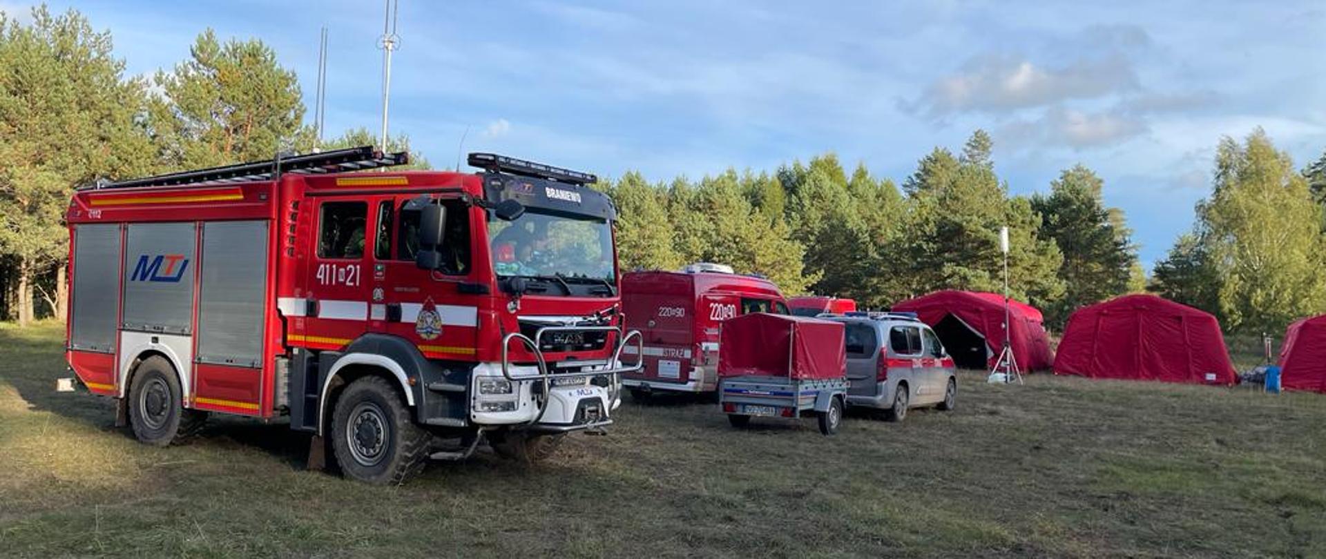 Na polanie ustawiony duży samochód strażacki. Z prawej strony czerwone namioty strażackie. W oddali drzewa i niebieskie niebo.