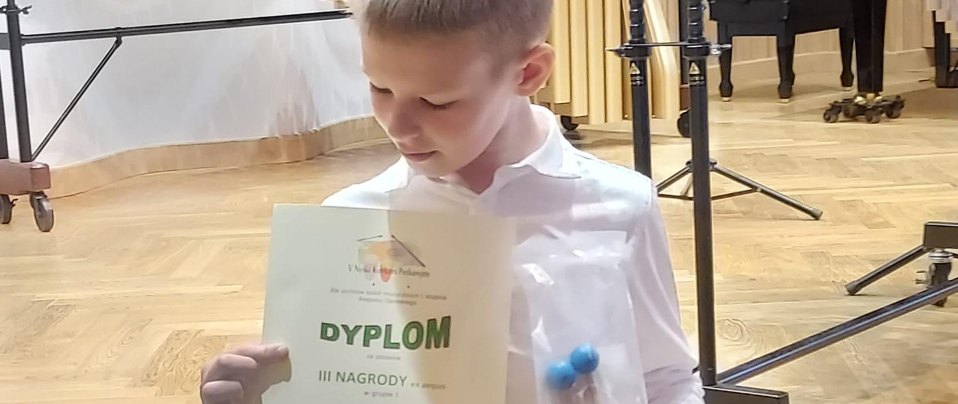 Uczeń w białej koszuli, trzymający w dłoni zdobyty dyplom, stojący przed sceną z jasnego drewna. Na scenie w tle instrumenty perkusyjne - marimba i wibrafon. W drugiej ręce trzyma pałki perkusyjne - z niebieskimi główkami.