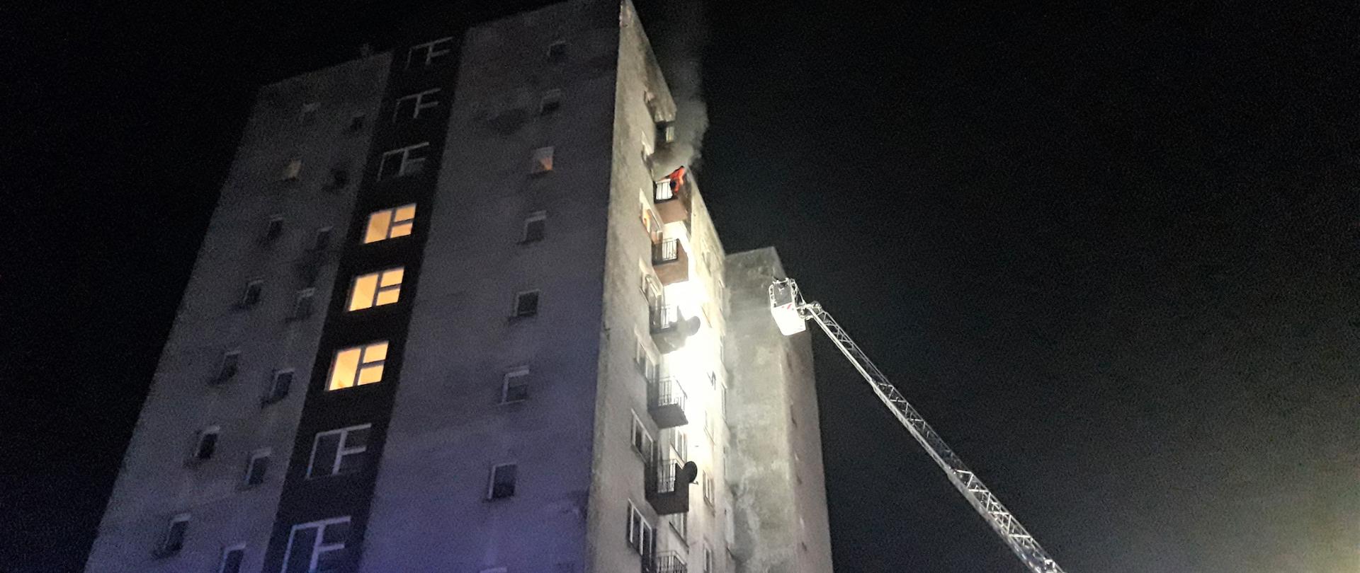 Fotografia przedstawia budynek wielorodzinny gdzie wybuchł pożar. Dym wydobywa się z okna balkonowego na zewnątrz, na balkonie stoi człowiek czekający na pomoc. Do ściany budynku zbliża się kosz drabiny mechanicznej.