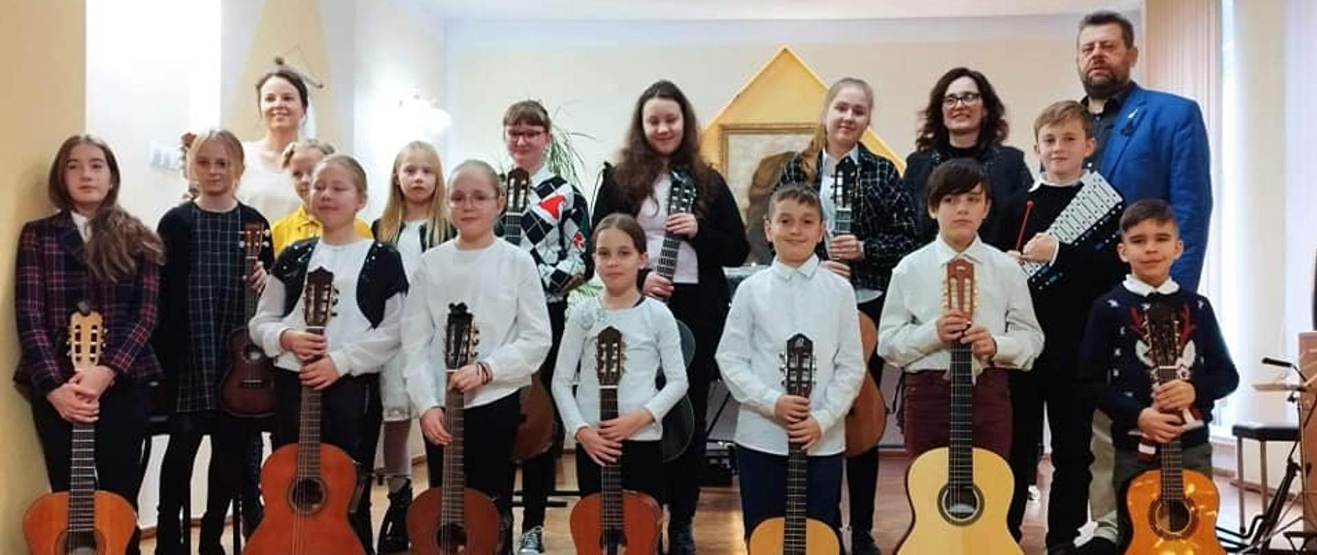 Zdjęcie przedstawia wszystkich wykonawców z koncertu " Kolędowy poranek gitarowy z dnia 17.12.2022 wraz z nauczycielami P. Agnieszką Kozakowską oraz P. Witoldem Kozakowskim. Zdjęcie wykonane w auli PSM I st. w Kłodzku, wszyscy uczniowie trzymają gitarę.