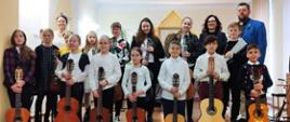 Zdjęcie przedstawia wszystkich wykonawców z koncertu " Kolędowy poranek gitarowy z dnia 17.12.2022 wraz z nauczycielami P. Agnieszką Kozakowską oraz P. Witoldem Kozakowskim. Zdjęcie wykonane w auli PSM I st. w Kłodzku, wszyscy uczniowie trzymają gitarę.