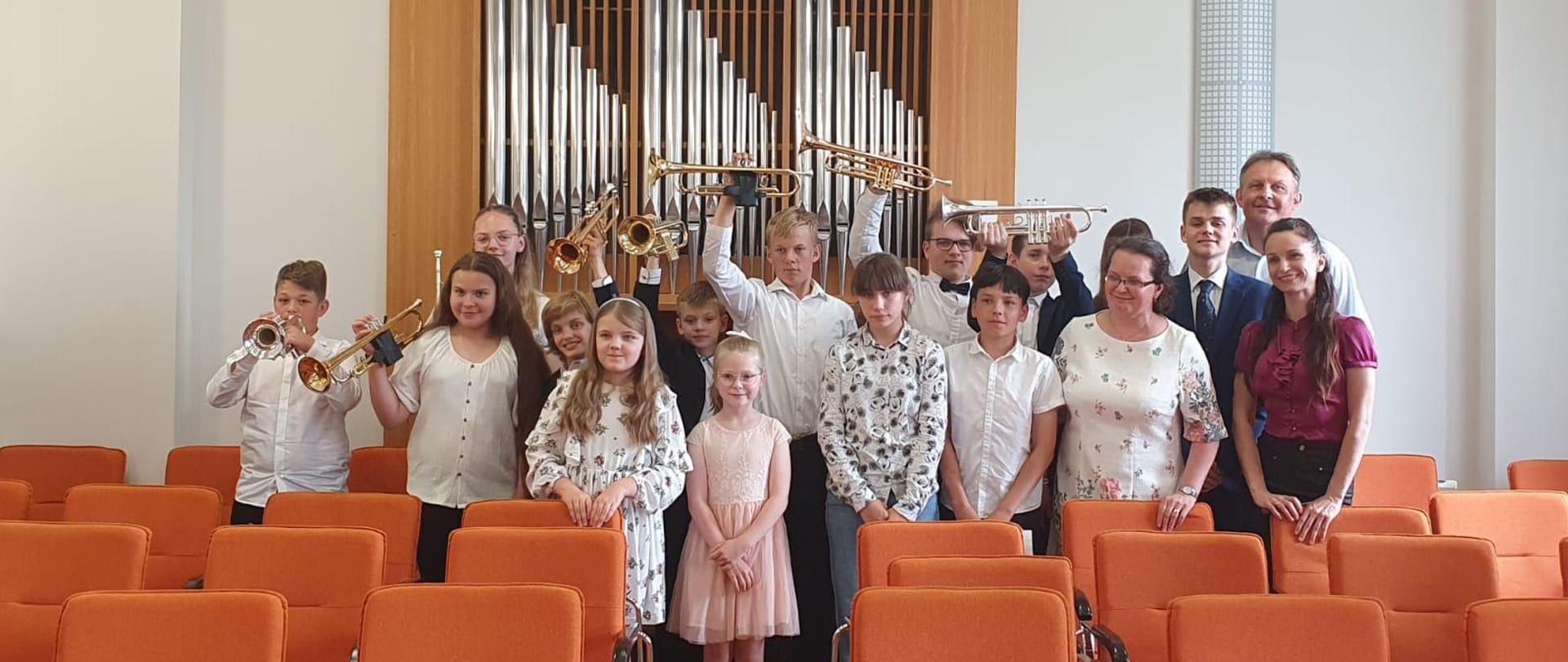 na zdjęciu grupa odświętnie ubranych uczniów. Część z nich trzyma podniesioną do góry trąbkę. W tyle za nimi piszczałki organowe. Przed grupą uczniów pomarańczowe krzesła.