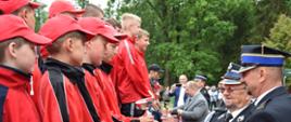 Chłopcy w czerwono czarnych strojach stoją na podium. Dwóch strażaków wręcza im puchar oraz medale. W tle mężczyzna który również wręcza puchar. Tło stanowią wysokie zielone drzewa. 