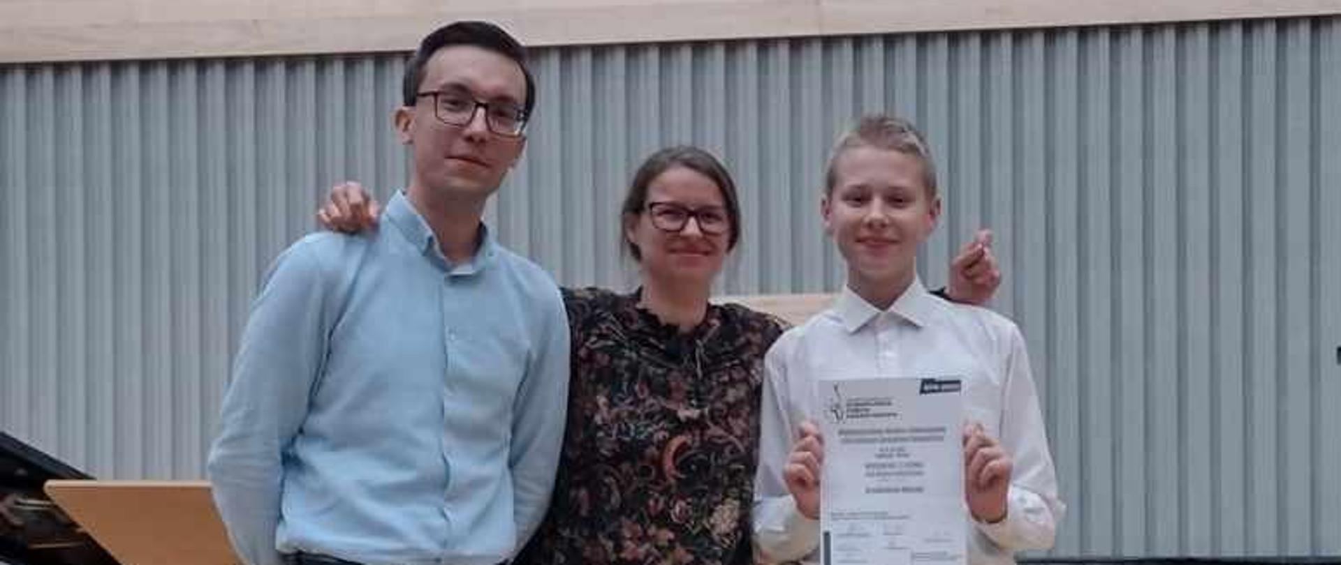 Uczeń trzymający dyplom wyróżnienia w towarzystwie nauczycieli na scenie.