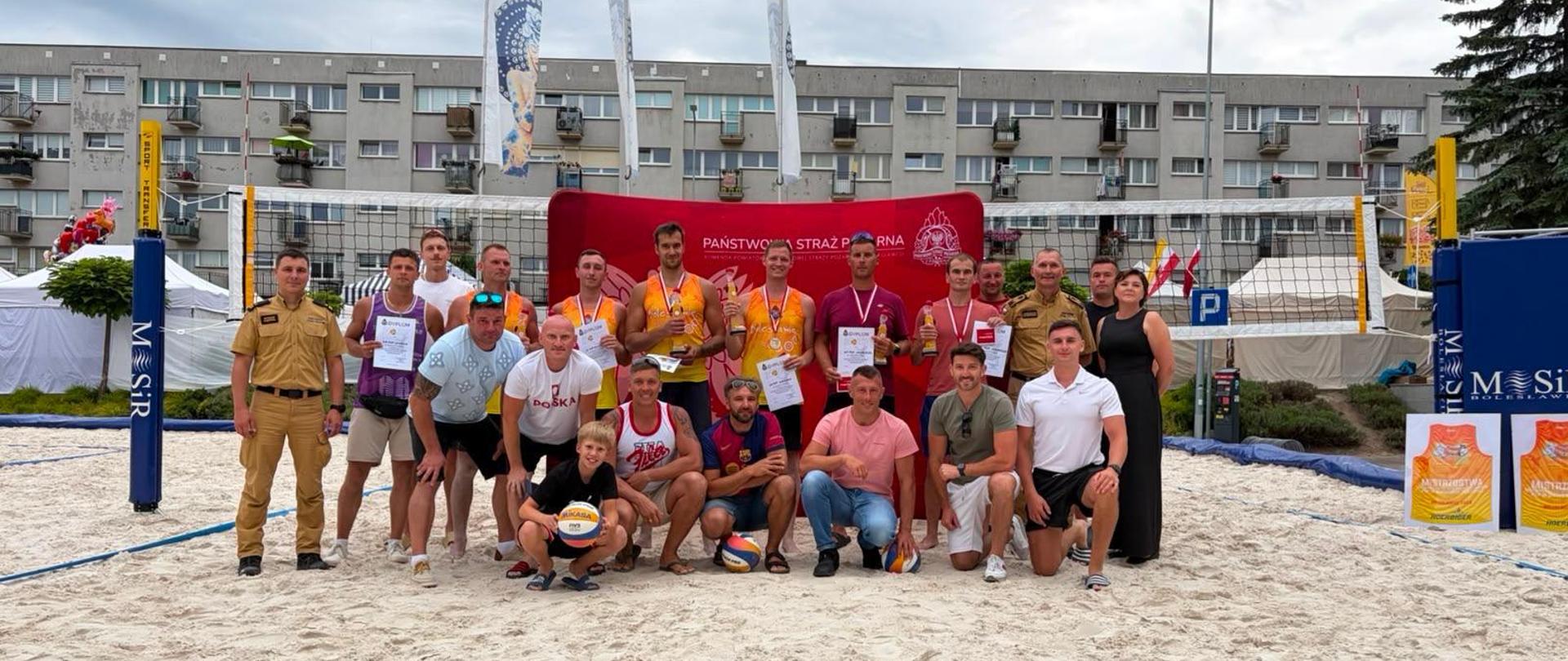 Pozujące na piasku duże pamiątkowe zdjęcie grupowe uczestników i organizatorów turnieju siatkówki plażowej. Część osób stoi z pucharami i dyplomami, a część kuca w przednim rzędzie. W tle znajduje się siatka do siatkówki, czerwona ścianka promocyjna straży pożarnej oraz flagi wydarzenia.