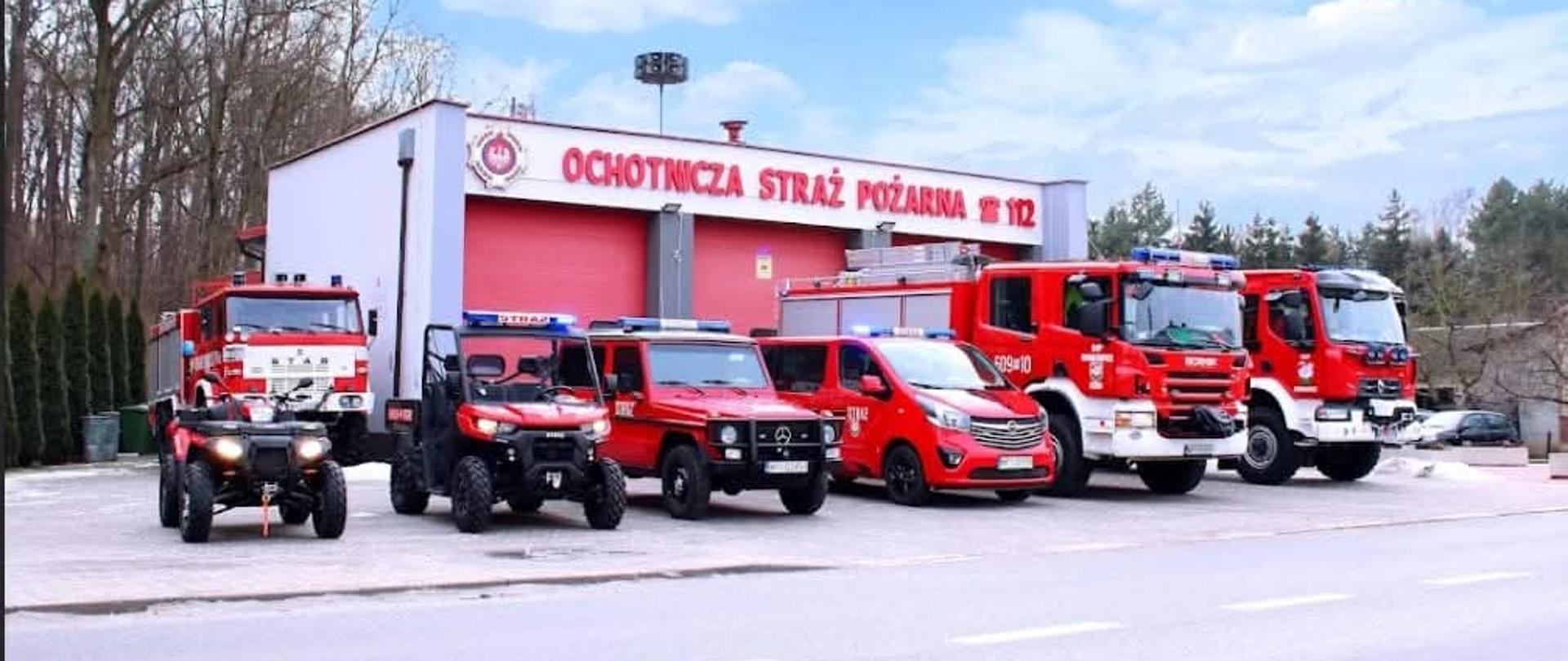 Zdjęcie przedstawia remizę Ochotniczej Straży Pożarnej (OSP) w Borkowicach, przed którą dumnie prezentuje się flota siedmiu pojazdów ratowniczych.
Budynek
W centralnej części znajduje się nowoczesny, biały budynek z trzema dużymi, czerwonymi bramami garażowymi. Nad bramami widnieje duży czerwony napis: "OCHOTNICZA STRAŻ POŻARNA" wraz z numerem alarmowym 112 oraz symbolem związku straży pożarnych.
Pojazdy (od lewej do prawej)
Flota jest bardzo zróżnicowana i przygotowana do różnych zadań terenowych oraz drogowych:
• Quad: Mały, czarny pojazd terenowy na grubych oponach.
• Star (ciężarowy): Klasyczny, czerwony wóz strażacki polskiej produkcji, zaparkowany nieco głębiej po lewej stronie.
• UTV (Side-by-Side): Lekki pojazd terenowy z klatką bezpieczeństwa i niebieskim oświetleniem ostrzegawczym.
• Mercedes-Benz klasy G: Terenowy samochód osobowy w barwach straży, idealny do trudnego terenu.
• Opel Vivaro (Bus): Czerwony furgon służący prawdopodobnie do transportu osób lub jako wóz kwatermistrzowski.
• Scania (ciężki wóz): Nowoczesny, masywny wóz gaśniczy z dużą kabiną załogową.
• Renault D14: Średni samochód ratowniczo-gaśniczy zamykający szereg po prawej stronie.
Otoczenie
Wszystkie pojazdy mają włączone niebieskie światła sygnalizacyjne (tzw. „koguty”). Akcja dzieje się w pogodny dzień przy lekkim zachmurzeniu. Po lewej stronie widać las, a przed remizą przebiega asfaltowa droga z wymalowanymi pasami. Na ziemi w niektórych miejscach widoczne są resztki śniegu.
