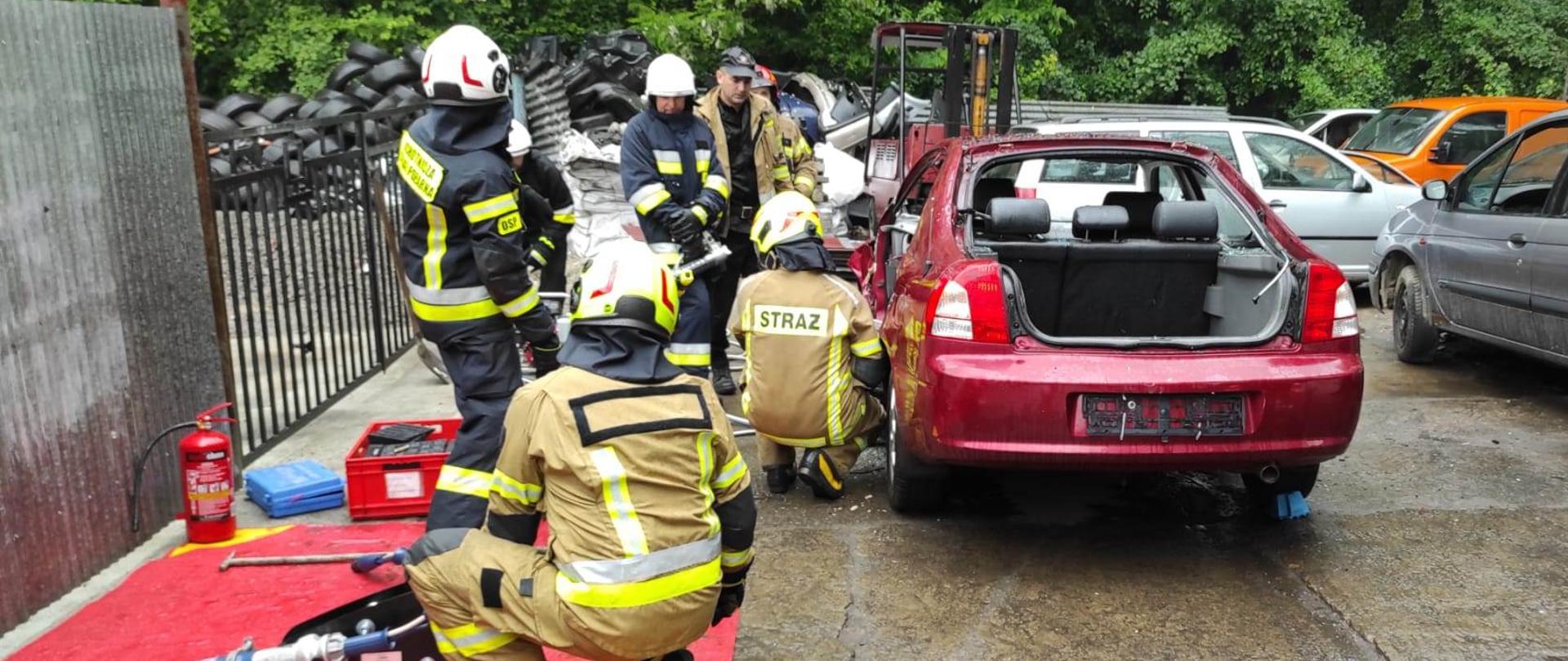 Kolorowe zdjęcie wykonane w pochmurny dzień, przedstawia utwardzony plac na którym widać kilku umundurowanych na bojowo strażaków. Ćwiczą ze sprzętem ratowniczym przy wraku pojazdu. Sprzęt przygotowany i zgromadzonych na czerwonej macie przy wraku. W tle widoczne inne drzewa i samochody. 