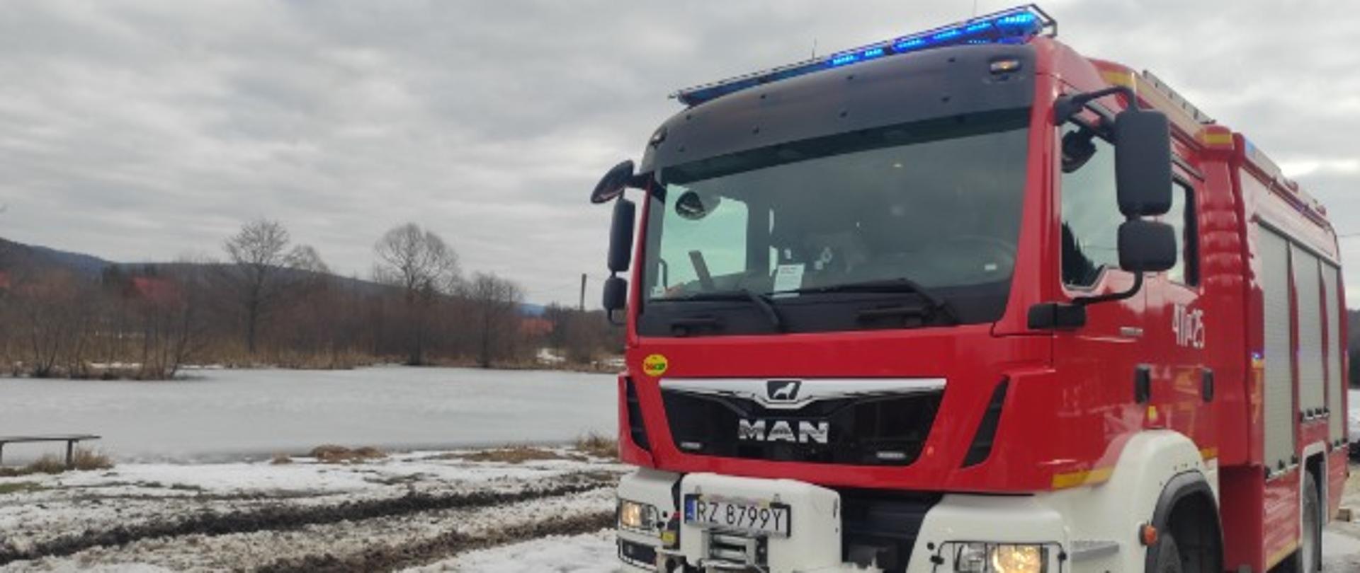 Kolorowa fotografia wykonana w pochmurny dzień. Przedstawia na pierwszym planie stojący czerwony pojazd ratowniczo-gaśniczy z włączonymi niebieskimi światłami alarmowymi. Teren pokryty jest śniegiem a w tle za pojazdem, widoczny jest zalodzony akwen. Daleko za akwenem widoczne szare drzewa. 