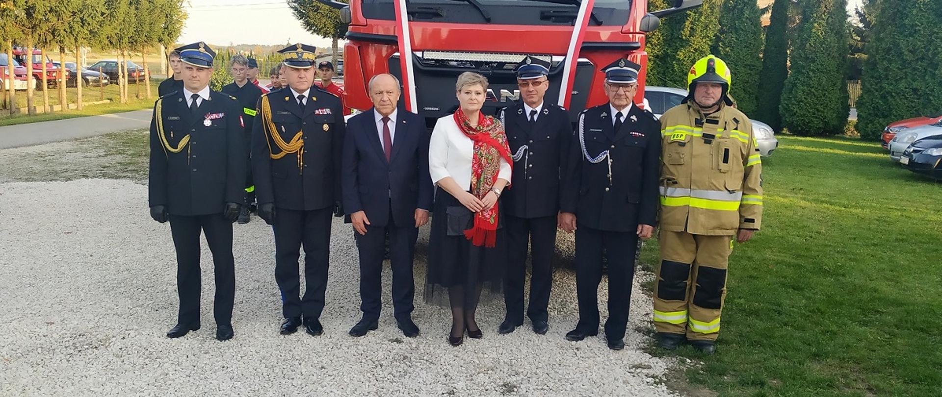 Fotografia grupowa upamiętniających gości na tle nowo przekazanego samochody ratowniczo-gaśniczego. Od prawej stoją ubrani w mundury galowe koloru granatowego Państwowej Straży Pożarnej: komendant powiatowy oraz podkarpacki komendant wojewódzki, następnie w środku: poseł na sejm RP oraz burmistrz miasta i gminy Przecław, a po lewej stronie druhowie jednostki OSP Tuszyma ubrani w galowe mundury koloru granatowego i ubranie bojowe typu nomex koloru piaskowego. 