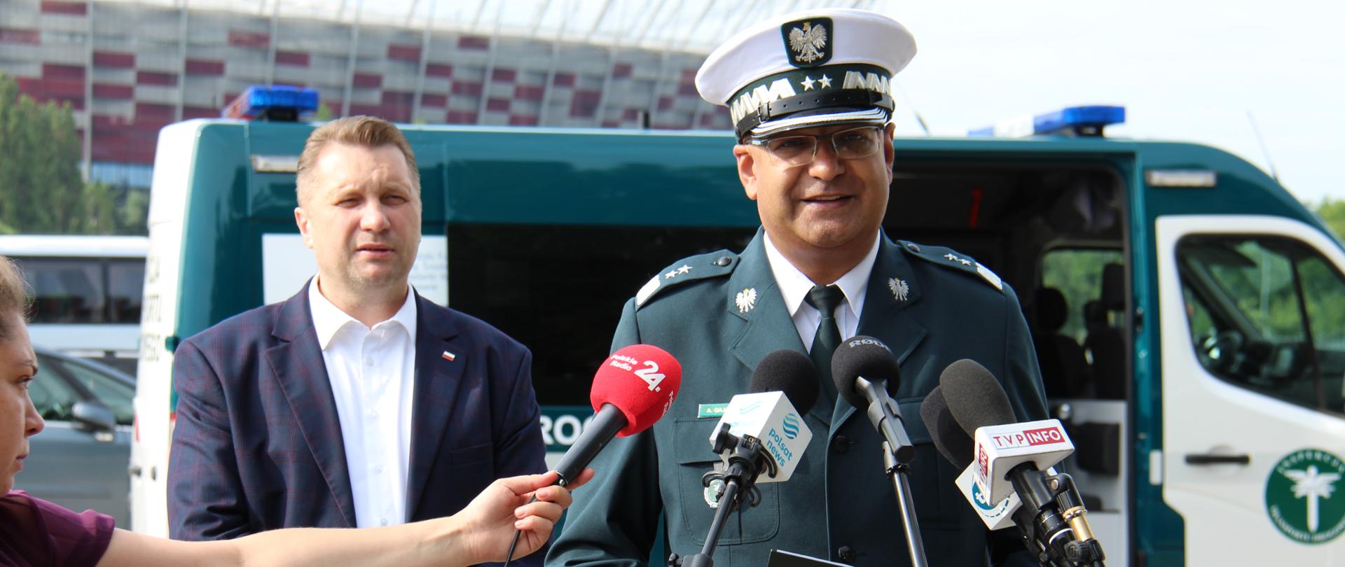 Na pierwszym tle (od lewej): Minister Edukacji i Nauki Przemysław Czarnek i Główny Inspektor Transportu Drogowego Alvin Gajadhur stoją przed mikrofonami. O rozpoczętych kontrolach autokarów turystycznych mówi Szef ITD. W tle: oznakowany furgon Inspekcji Transportu Drogowego i Stadion Narodowy w Warszawie.