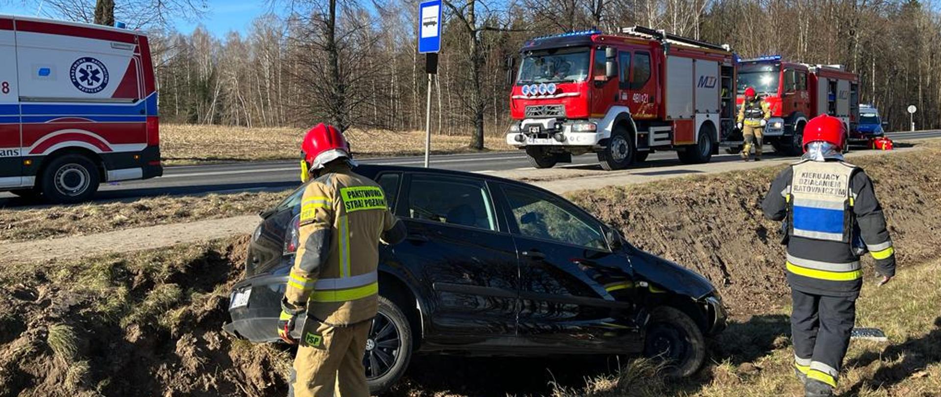 Zdjęcie przedstawia uszkodzony samochód znajdujący się w przydrożnym rowie oraz strażaków PSP