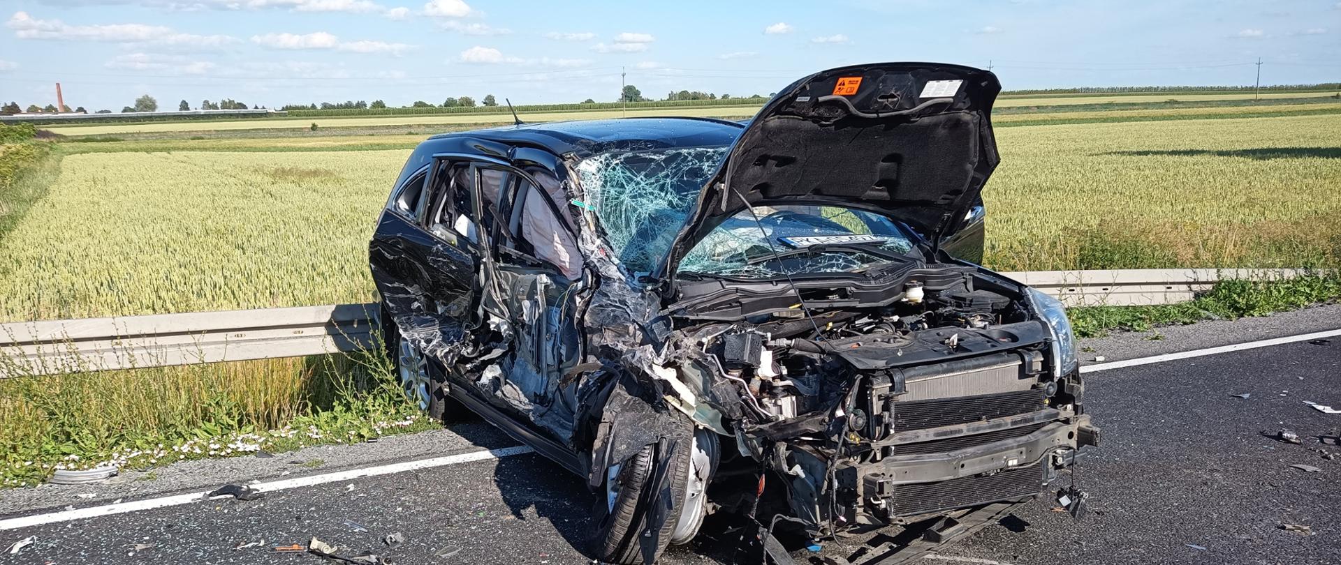 Na zdjęciu widać uszkodzony pojazd. Droga asfaltowa. Po bokach pola uprawne. Niebo błękitne. Lekkie zachmurzenie. Na asfalcie fragmenty karoserii. Metalowa bariera drogowa. 