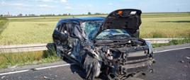 Na zdjęciu widać uszkodzony pojazd. Droga asfaltowa. Po bokach pola uprawne. Niebo błękitne. Lekkie zachmurzenie. Na asfalcie fragmenty karoserii. Metalowa bariera drogowa. 