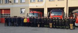 Zdjęcie przedstawia funkcjonariuszy i emerytowanych strażaków na tle budynku Jednostki Ratowniczo-Gaśniczej w Krotoszynie oraz samochodów pożarniczych podczas wspólnego zdjęcia.
