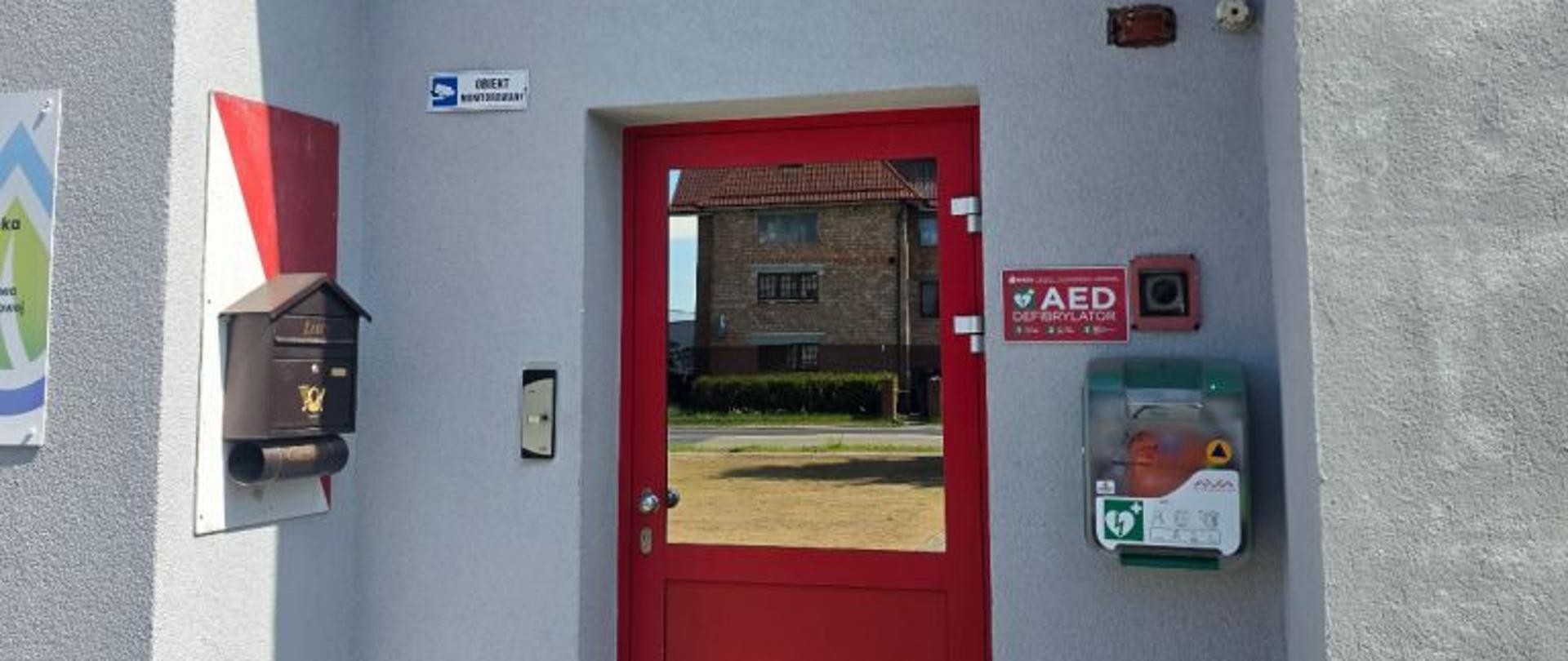 Pionowe zdjęcie przedstawiające główne wejście do budynku wykonanego z jasnoszarego, strukturalnego tynku. W centralnej części kadru znajdują się jednoskrzydłowe, przeszklone drzwi wejściowe w wyrazistej, czerwonej ramie, w których szybie odbija się sąsiedni budynek z cegły oraz bezchmurne niebo. Nad drzwiami zamontowane są dwa elementy identyfikacyjne: owalne godło Polski z Białym Orłem na czerwonym tle, a bezpośrednio pod nim prostokątna, czerwona tablica urzędowa z białym napisem „KOMENDA POWIATOWA PAŃSTWOWEJ STRAŻY POŻARNEJ w Białogardzie woj. zachodniopomorskie”. Po prawej stronie drzwi, na wysokości wzroku, zamontowana jest zielono-szara, plastikowa skrzynka defibrylatora AED marki Aivia z widocznym przez przezroczystą pokrywą pomarańczowym urządzeniem, a nad nią wisi czerwona tabliczka informacyjna z białym napisem „AED DEFIBRYLATOR” oraz czarny włącznik w czerwonej obudowie. Po lewej stronie drzwi widoczna jest brązowa, metalowa skrzynka na listy, fragment biało-czerwonego malowania pasowego na ścianie oraz mała, niebiesko-biała tabliczka z napisem „OBIEKT MONITOROWANY”. Przed wejściem znajduje się podłoże wyłożone szarą kostką brukową, na którą pada wyraźny cień budynku.