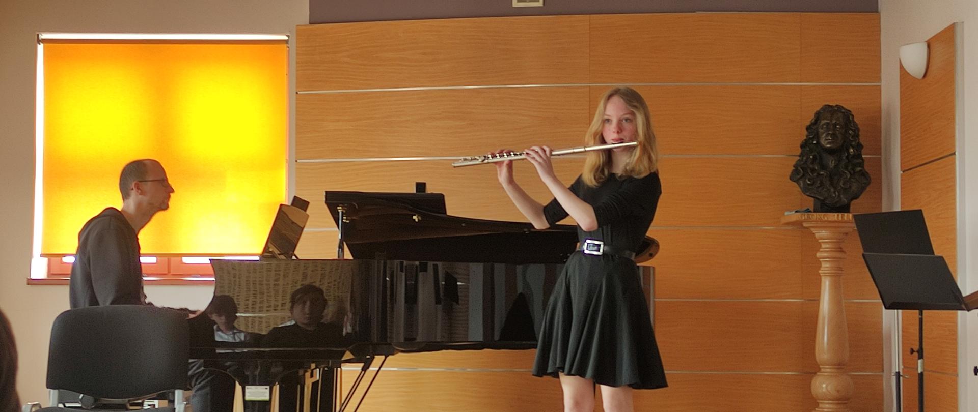 Na scenie sali kameralnej uczennica gra na flecie, za nią trzy pulpity pulpity. Na pierwszym planie puste krzesła. Z lewej strony fortepian, przy którym gra akompaniatorka zerkając na nuty. Z tyłu okno przykryte roletą